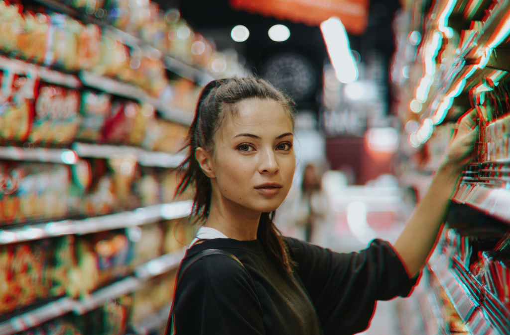 A woman facing the camera in a supermarket aisle. She is reaching for a product on the shelf.